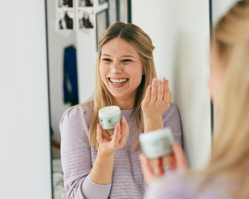Femme tenant un pot de crème jour Formy et s'appliquant de la crème sur le nez en souriant dans le miroir