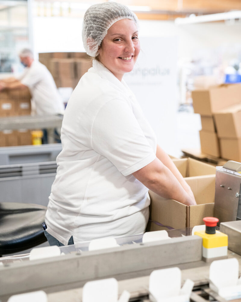 Femme avec une charlotte sur la tête mettant des produits dans une boîte en carton sur une ligne de production.